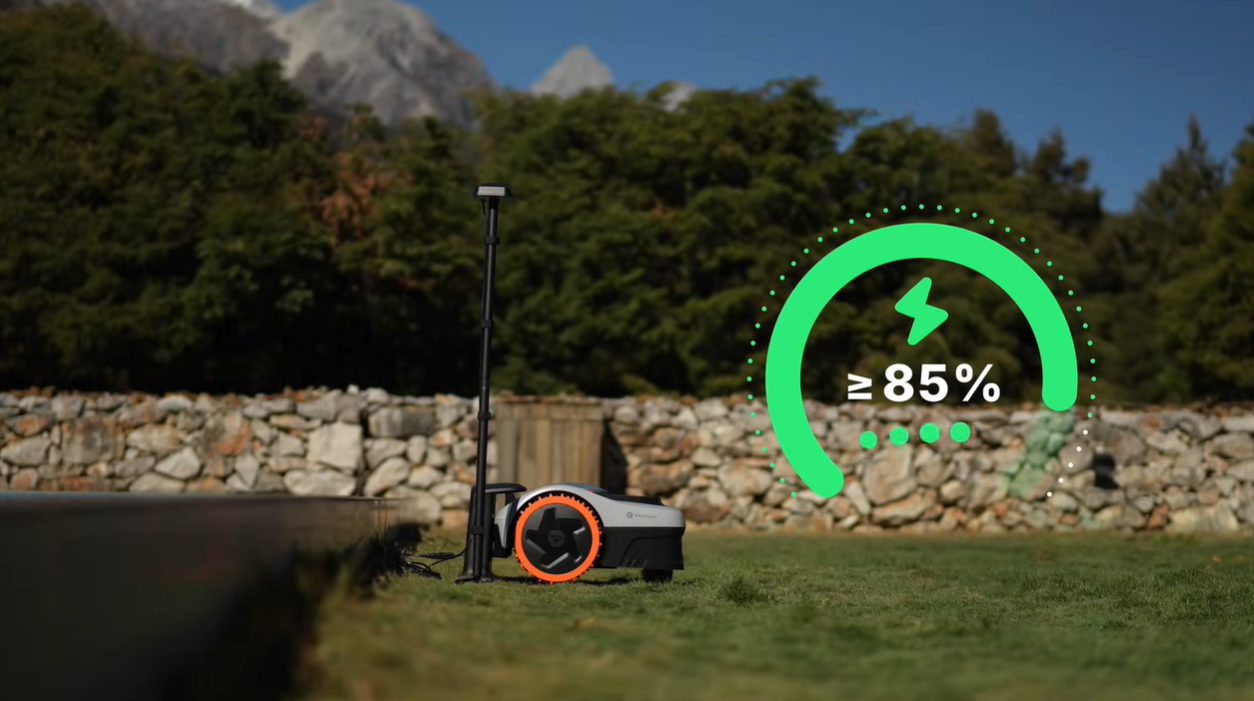 En robotgräsklippare parkerad i sin laddstation utomhus tillsammans med en grön grafik som poängterar vikten av att ladda batteriet till minst 85 procent innan vintervilan.