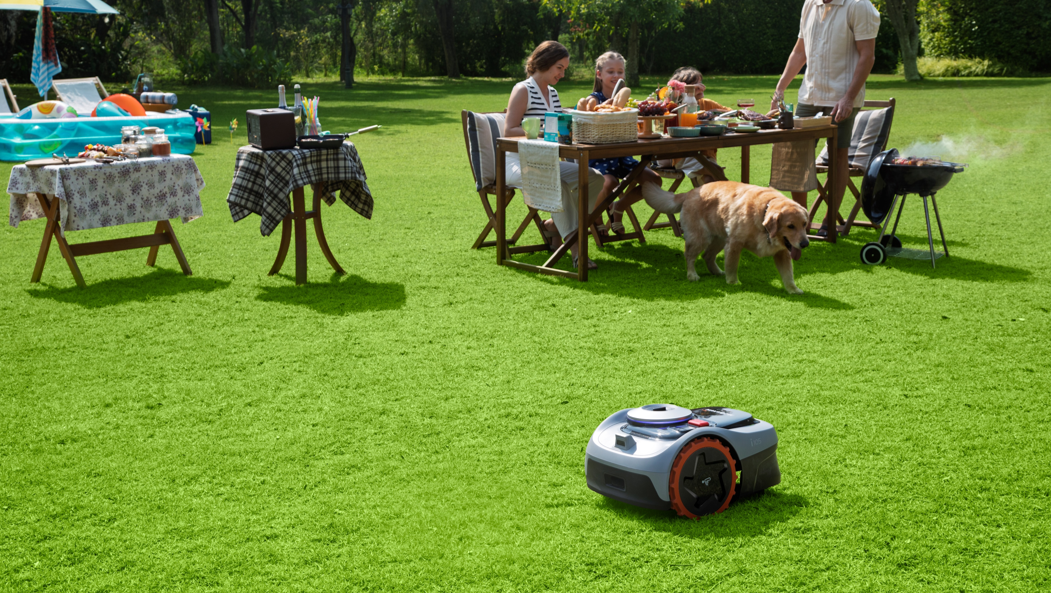 En modern slinglös robotgräsklippare arbetar tyst och säkert på den perfekta gräsmattan medan en familj och deras hund njuter av en grillfest i bakgrunden.